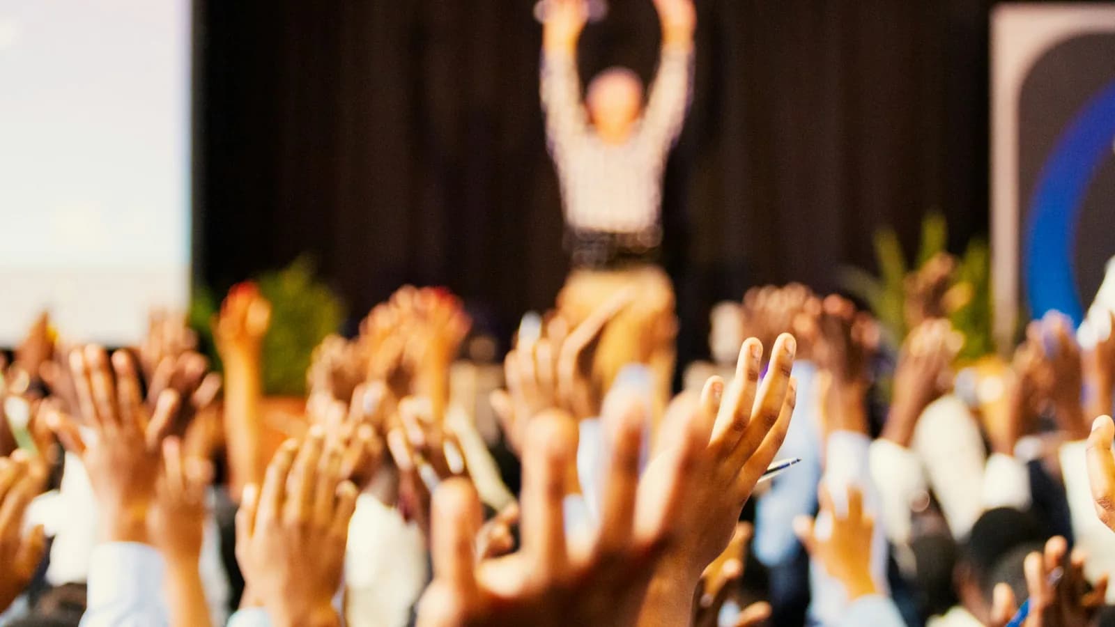 Audience with raised hands at a live event, speaker lit on stage behind them